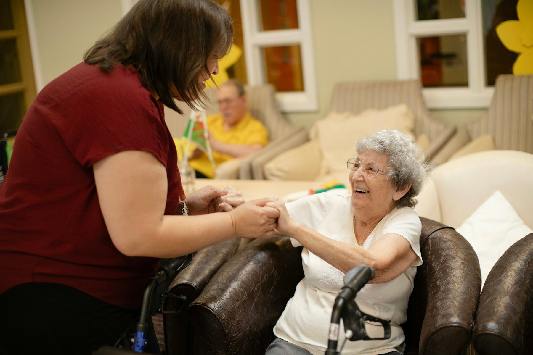 A caregiver holds the hands of a smiling older woman with a walker, helping her stand from her chair.