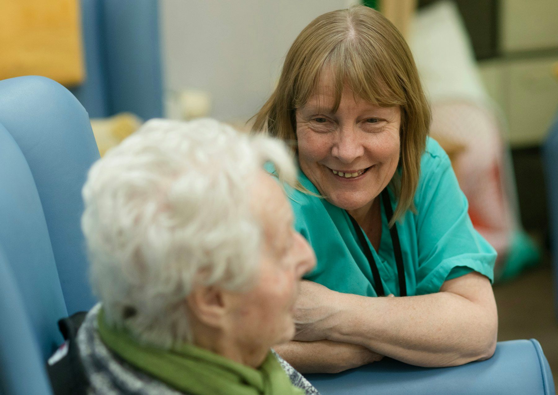 A care professional in teal scrubs smiles warmly while talking with an older woman seated in an armchair.